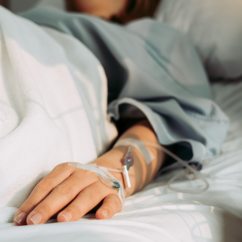 Eine soeben operierte Person liegt im Krankenbett, im Fokus ist eine angeschlossene Infusion.