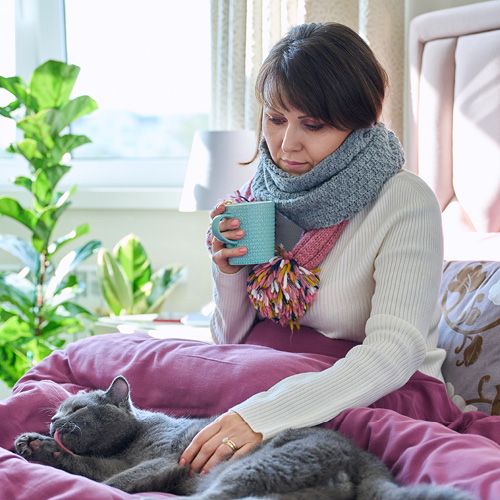 Eine erkältete Frau sitzt mit Schal und Tee im Bett, während sie eine Katze streichet.