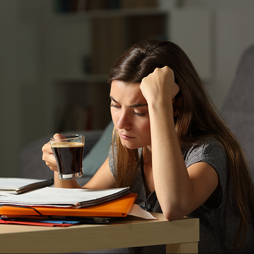 Eine Studentin hängt mit einem frischen Kaffee in der Hand verzweifelt über ihren Lernzetteln.